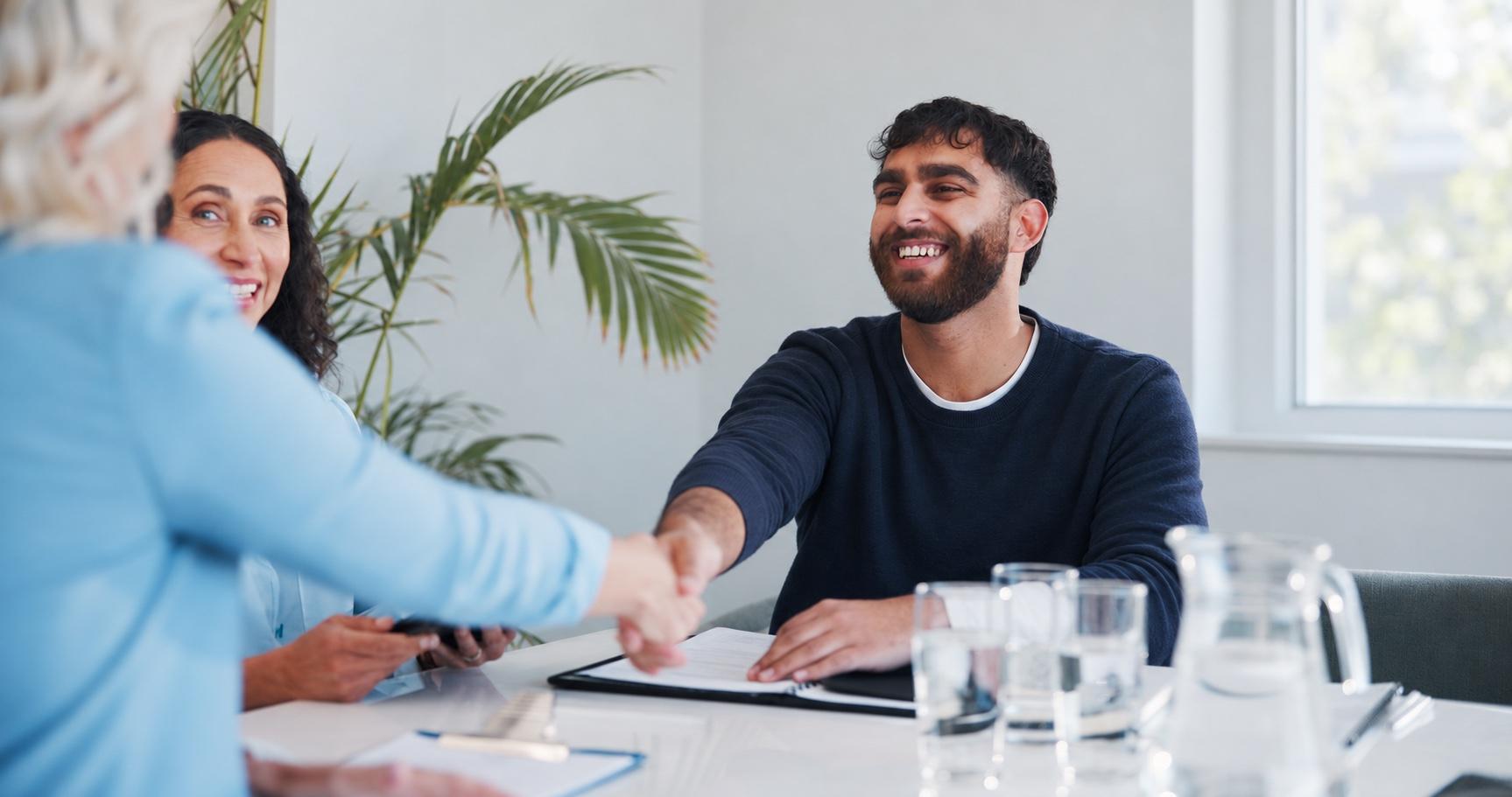 Ein Bewerber und eine Gesprächspartnerin reichen sich im Vorstellungsgespräch die Hand, daneben eine weitere Gesprächsteilnehmerin.